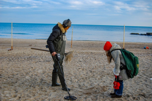 Two people on a beach. A male using a metal detector. The female is observing.