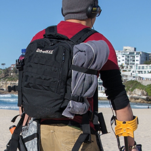 A man metal detecting out on the beach wearing a back pack.